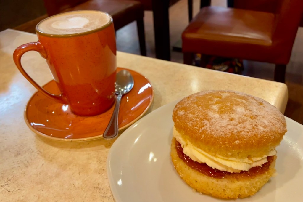 A hot cup of cappuccino in an orange mug on a saucer with a teaspoon, alongside a plate of sponge cake filled with cream and jam.